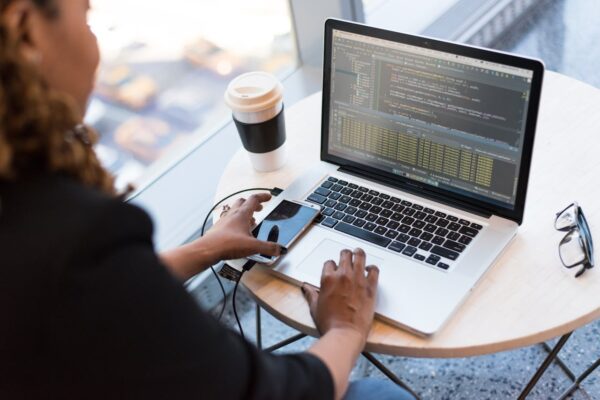 Black woman programming on a laptop with coffee, smartphone, and glasses on a desk in an office.