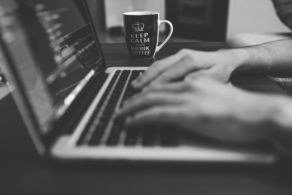 Monochrome image of hands on a laptop keyboard with a coffee mug, coding in progress.