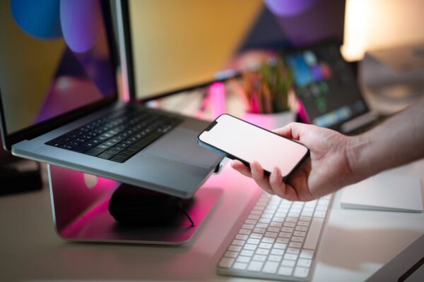 Close-up of a hand holding a smartphone with blank screen next to a laptop in a modern office.